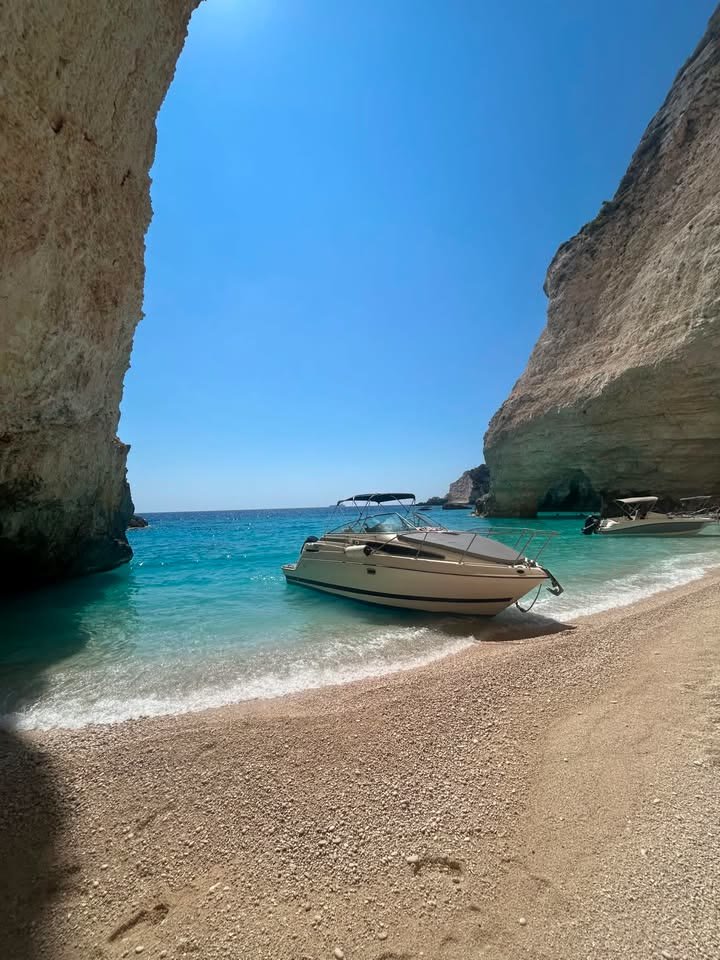 ΝΙΚΟΛΑΟΣ pulled up on a narrow pebble beach seen through a gap between two towering white cliffs, with a second boat further along the shore