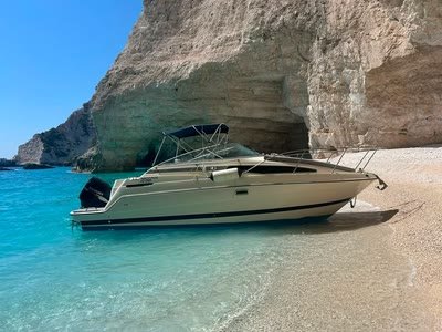 ΝΙΚΟΛΑΟΣ beached on pebbles directly beneath a huge pale cliff, turquoise shallows in front