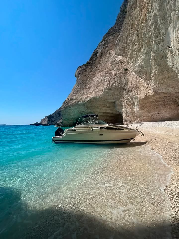 The boat resting on the shoreline of a deserted cove directly beneath a huge overhanging pale cliff, the water shading from clear shallows to deep turquoise
