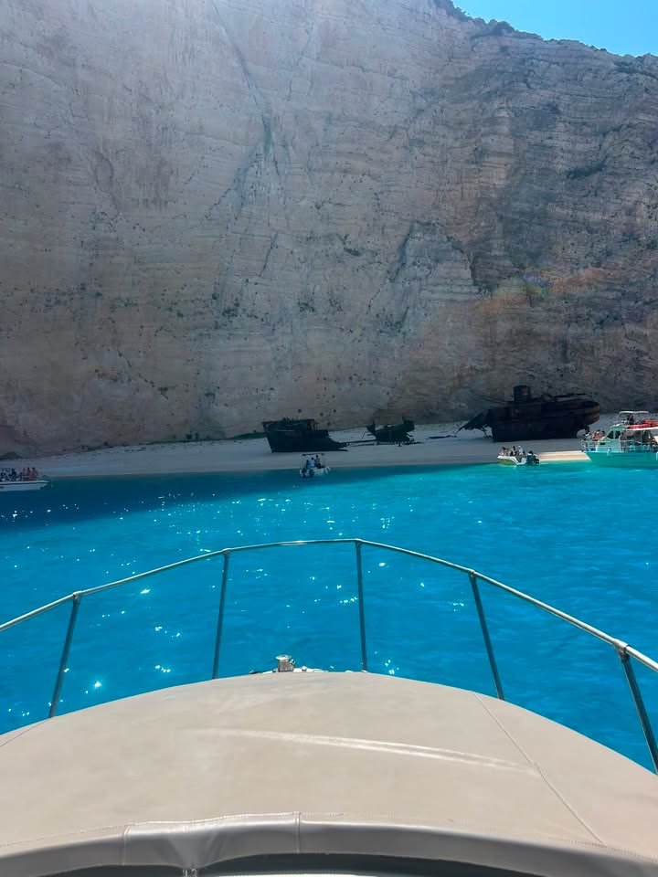 The rusted shipwreck on the white sand of Navagio beach beneath a towering pale cliff, brilliant turquoise water in front, seen over the bow of the MareWaves boat